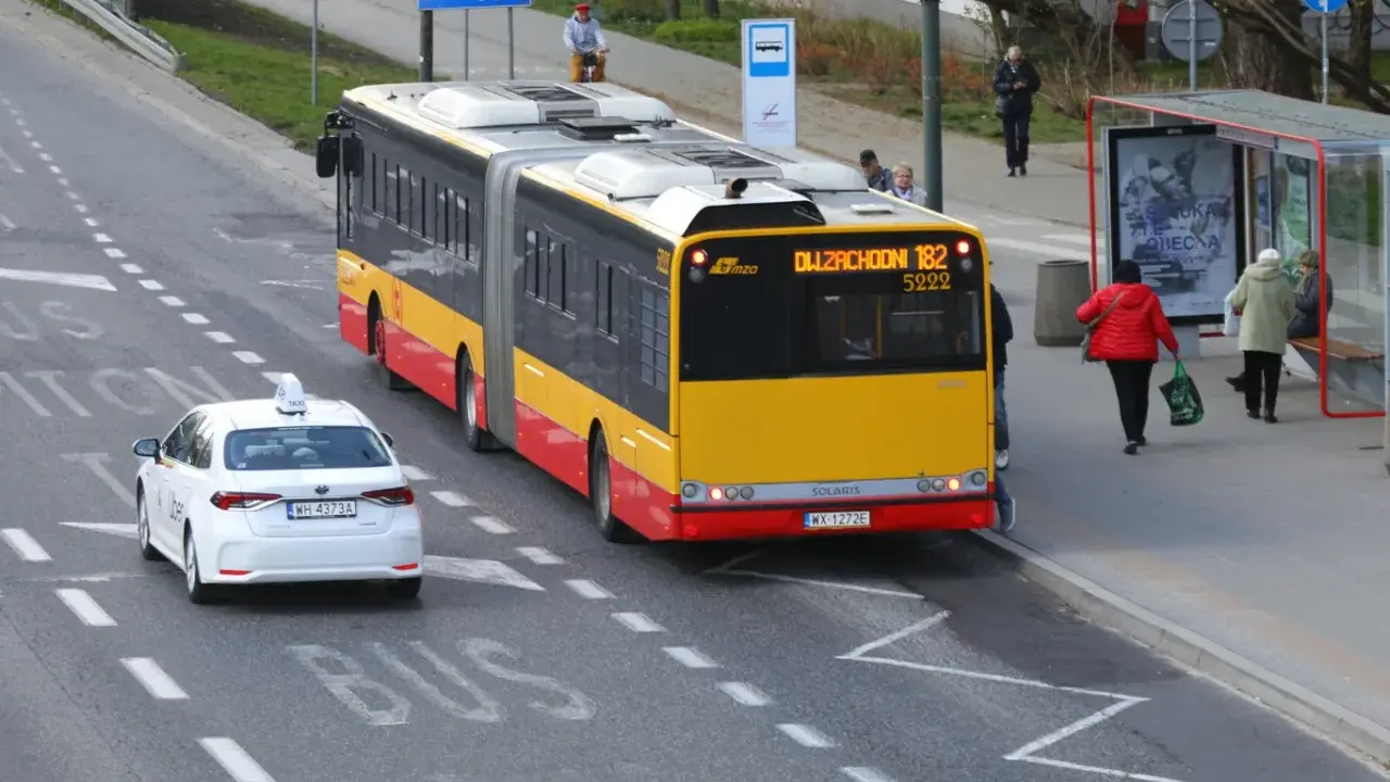 Autobus linii 182 zbliża się do przystanku, a biała taksówka czeka, by ustąpić mu pierwszeństwa.