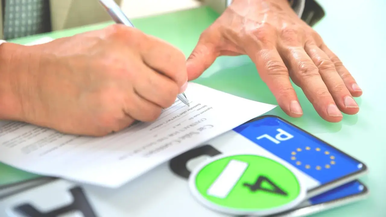 Podpisanie umowy kupna-sprzedaży pojazdu, co jest kluczowe dla uzyskania homologacji. Obok dokumentów leżą tablice rejestracyjne.