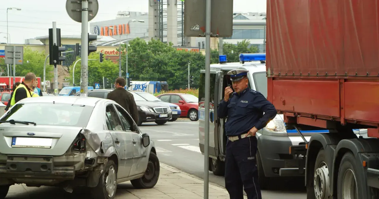 Policjant rozmawia przez radio przy miejscu wypadku. Uszkodzone auto i ciężarówka sugerują, że mogło dojść do zdarzenia związanego z pzu klauzula alkoholowa.