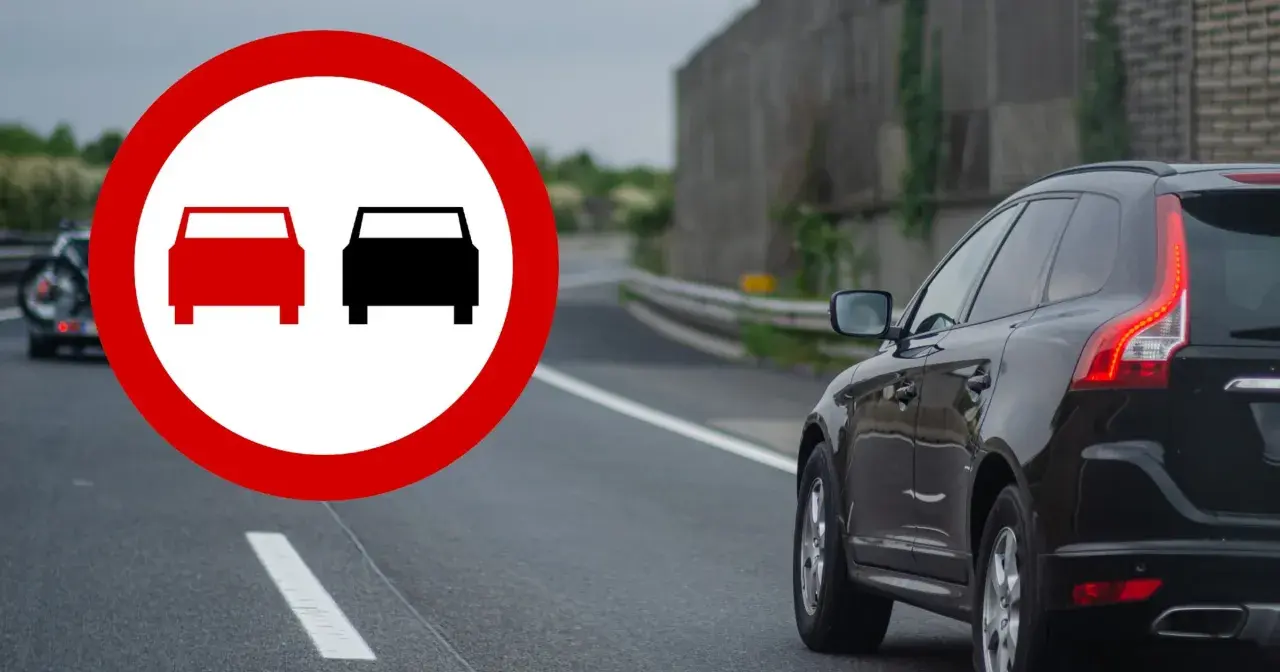 Znak zakazu wyprzedzania na podwójnej ciągłej. Czarny SUV jedzie po drodze, a na pierwszym planie znak ostrzegawczy.