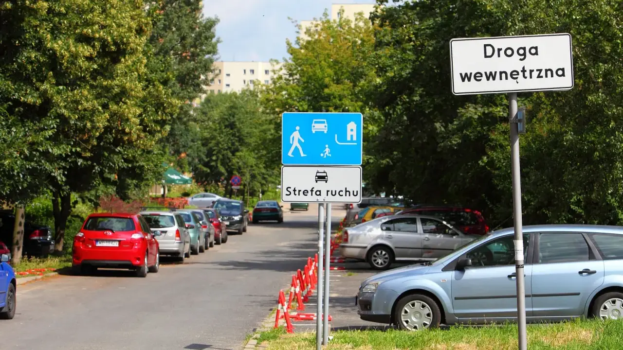 Znak "Droga wewnętrzna" obok strefy ruchu z piktogramami pieszych i samochodów. Zaparkowane samochody wzdłuż ulicy.