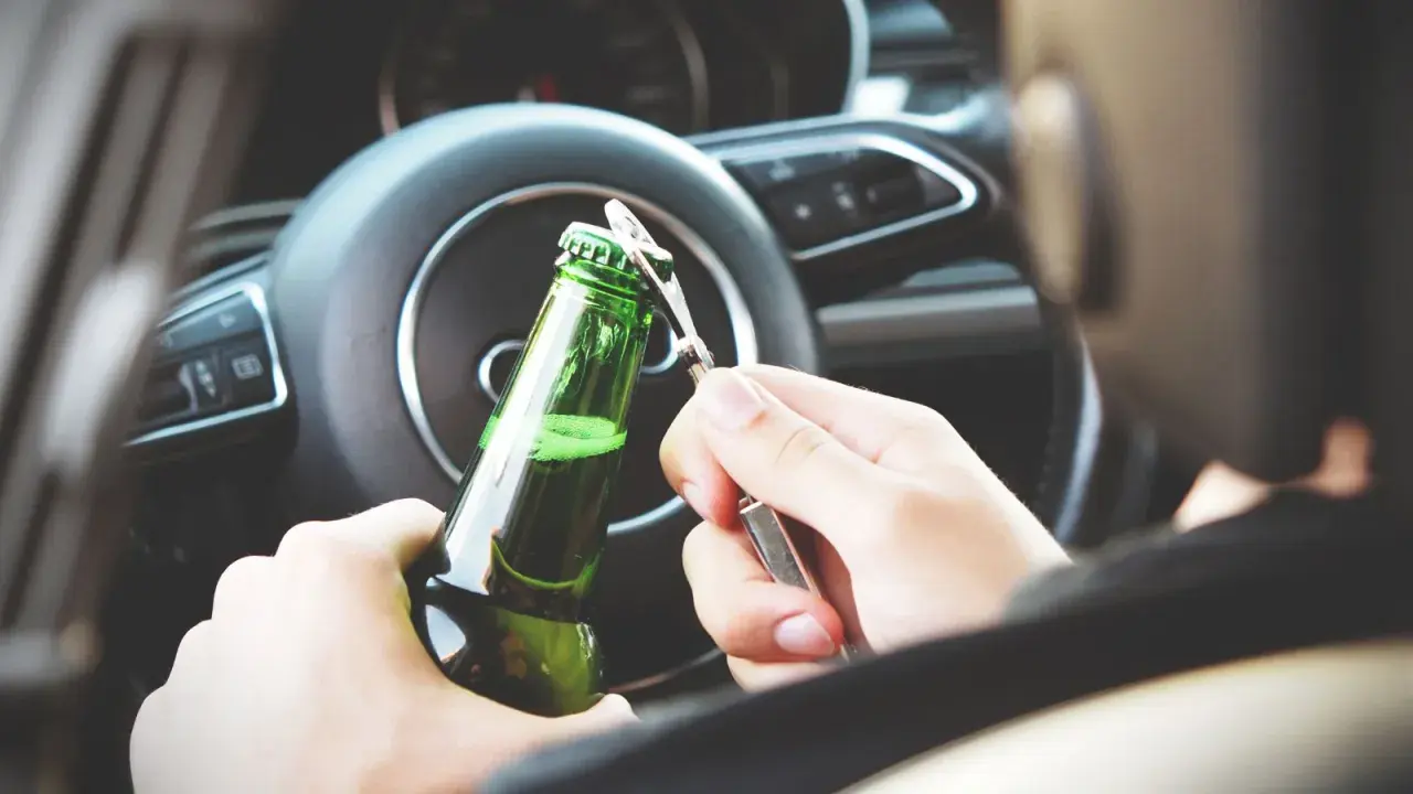 Ręce otwierające butelkę piwa za kierownicą. Pamiętaj, że od 0,2 promila zabierają auto.