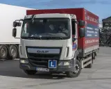 Biała ciężarówka DAF z naczepą, na której widnieje napis "Kategoria C1". Za kierownicą siedzi kierowca w masce.