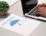 Osoba pisze na laptopie, obok dokument z napisem "INSURANCE" i parasolką. Szuka najlepszych znizki na ubezpieczenie auta.