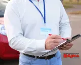 Instruktor notuje dopuszczalne błędy na egzaminie praktycznym na prawo jazdy. Obok czerwony samochód.