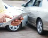 Mężczyzna z tabletem w rękach, obok dwa samochody. Sprawdza ubezpieczenie samochodu firmowego po kolizji.