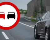 Znak zakazu wyprzedzania na podwójnej ciągłej. Czarny SUV jedzie po drodze, a na pierwszym planie znak ostrzegawczy.