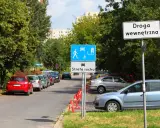 Znak "Droga wewnętrzna" obok strefy ruchu z piktogramami pieszych i samochodów. Zaparkowane samochody wzdłuż ulicy.