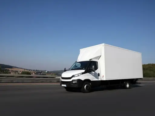 Biały samochód ciężarowy powyżej 3,5 tony pędzi autostradą. W tle krajobraz z pagórkami i domami.