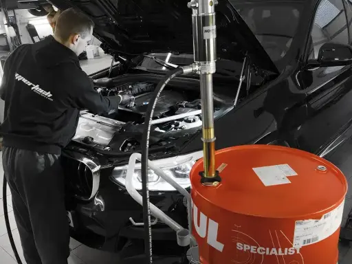 Mechanik wymienia olej w czarnym BMW. Duży, pomarańczowy bęben z napisem "Motul" sugeruje, że to czas na regularną wymianę oleju.