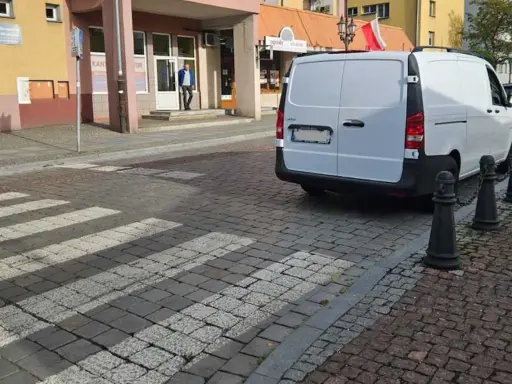 Biały van blokuje przejście dla pieszych na brukowanej ulicy.