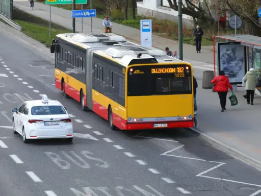 Autobus linii 182 zbliża się do przystanku, a biała taksówka czeka, by ustąpić mu pierwszeństwa.