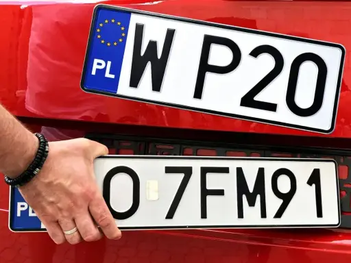 Dłoń z bransoletką montuje tablice rejestracyjne na czerwonym samochodzie. Widać dwie tablice: jedną z napisem WP20, drugą z numerem O-7FM91.