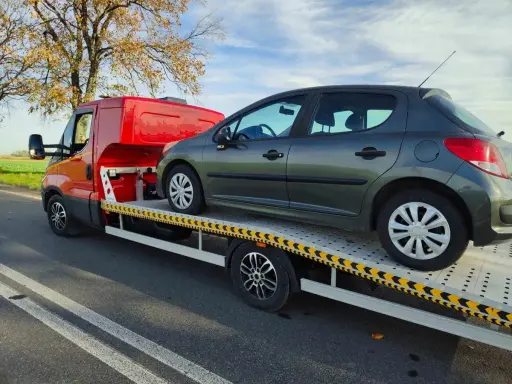 Ciemnoszary samochód na lawecie, przygotowany do konfiskaty. Pomarańczowy pojazd pomocy drogowej stoi na poboczu drogi.