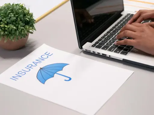 Osoba pisze na laptopie, obok dokument z napisem "INSURANCE" i parasolką. Szuka najlepszych znizki na ubezpieczenie auta.