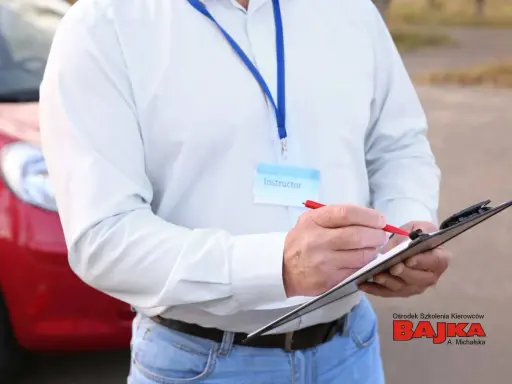 Instruktor notuje dopuszczalne błędy na egzaminie praktycznym na prawo jazdy. Obok czerwony samochód.