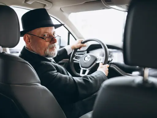 Starszy pan w kapeluszu i okularach prowadzi samochód. W ręku trzyma dowód rejestracyjny, który jest dowodem współwłaściciel.