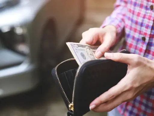Ręce wyciągają banknot 50 dolarów z portfela. W tle widać fragment samochodu, sugerując kupno samochodu za gotówkę.