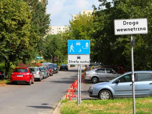 Znak "Droga wewnętrzna" obok strefy ruchu z piktogramami pieszych i samochodów. Zaparkowane samochody wzdłuż ulicy.