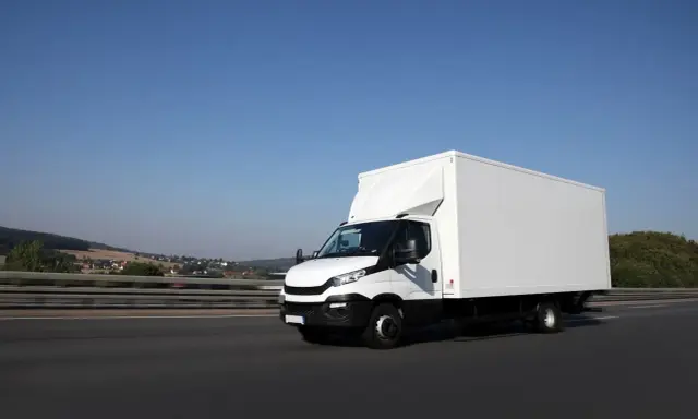 Biały samochód ciężarowy powyżej 3,5 tony pędzi autostradą. W tle krajobraz z pagórkami i domami.
