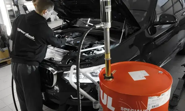 Mechanik wymienia olej w czarnym BMW. Duży, pomarańczowy bęben z napisem "Motul" sugeruje, że to czas na regularną wymianę oleju.