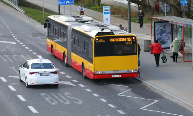 Autobus linii 182 zbliża się do przystanku, a biała taksówka czeka, by ustąpić mu pierwszeństwa.