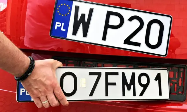 Dłoń z bransoletką montuje tablice rejestracyjne na czerwonym samochodzie. Widać dwie tablice: jedną z napisem WP20, drugą z numerem O-7FM91.