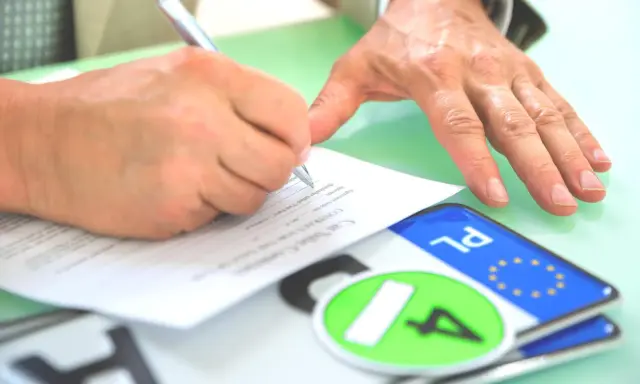 Podpisanie umowy kupna-sprzedaży pojazdu, co jest kluczowe dla uzyskania homologacji. Obok dokumentów leżą tablice rejestracyjne.