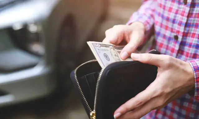 Ręce wyciągają banknot 50 dolarów z portfela. W tle widać fragment samochodu, sugerując kupno samochodu za gotówkę.