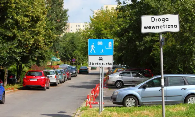 Znak "Droga wewnętrzna" obok strefy ruchu z piktogramami pieszych i samochodów. Zaparkowane samochody wzdłuż ulicy.
