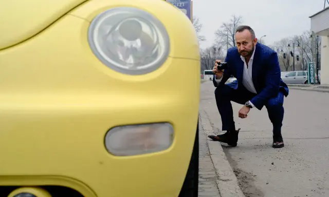 Rzeczoznawca samochodowy w garniturze klęczy przy żółtym samochodzie, robiąc zdjęcie aparatem.