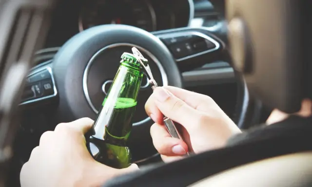 Ręce otwierające butelkę piwa za kierownicą. Pamiętaj, że od 0,2 promila zabierają auto.