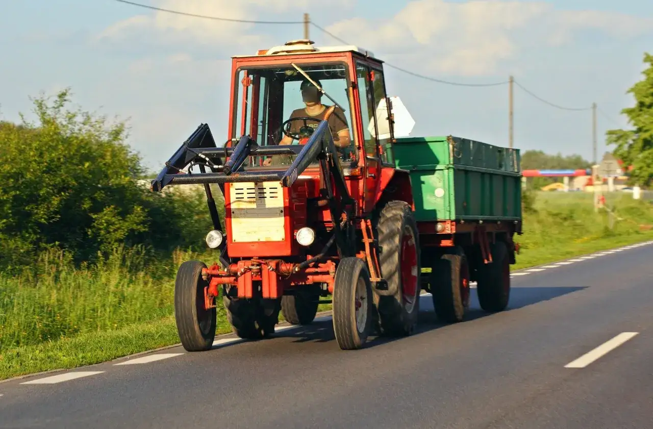 Czerwony ciągnik z przyczepą jedzie drogą. Kierowca w kabinie, zielona skrzynia ładunkowa. Przegląd ciągnika w pełnym słońcu.