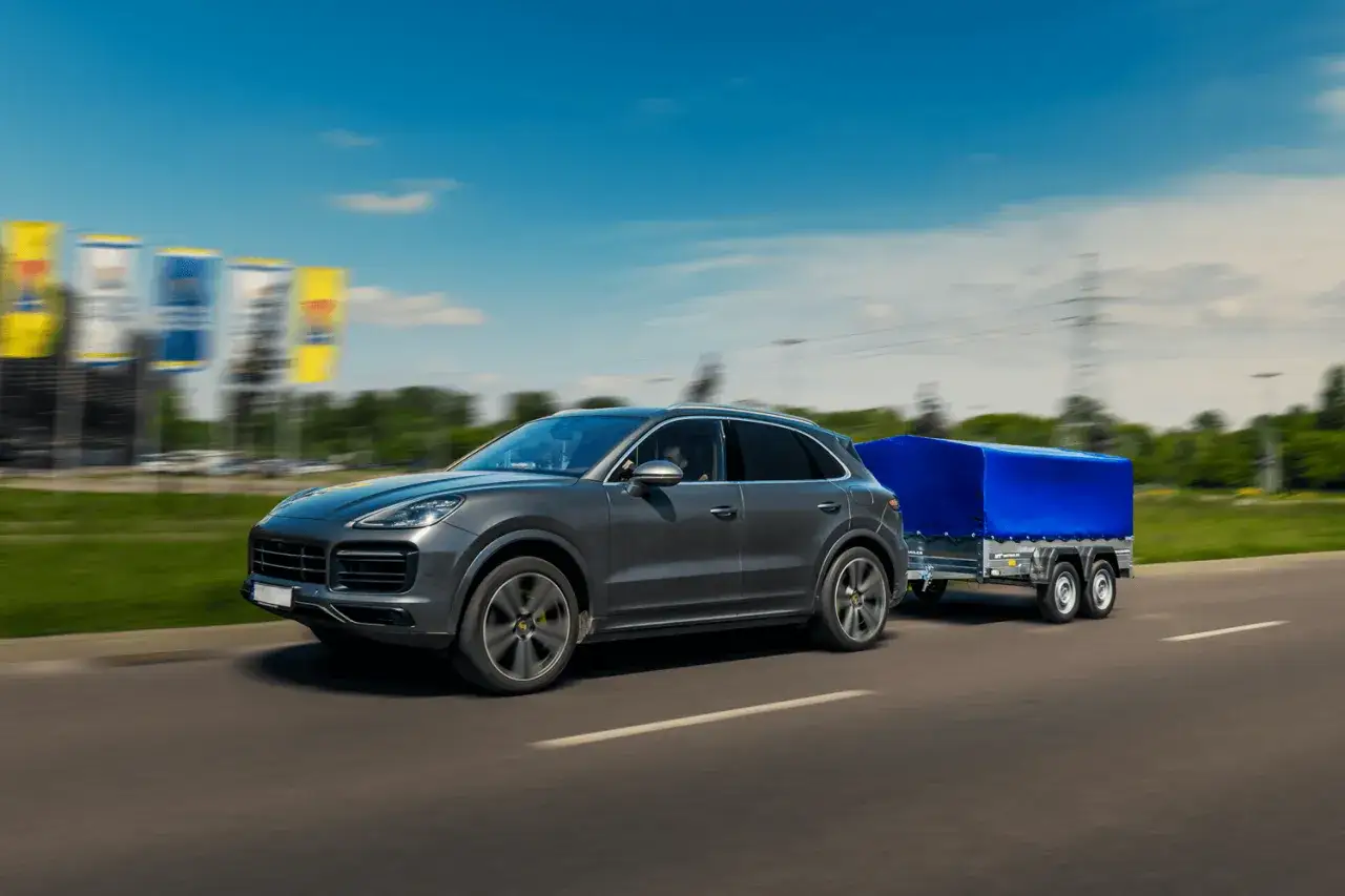 Szary SUV z przyczepką pod niebieską plandeką jedzie po drodze.