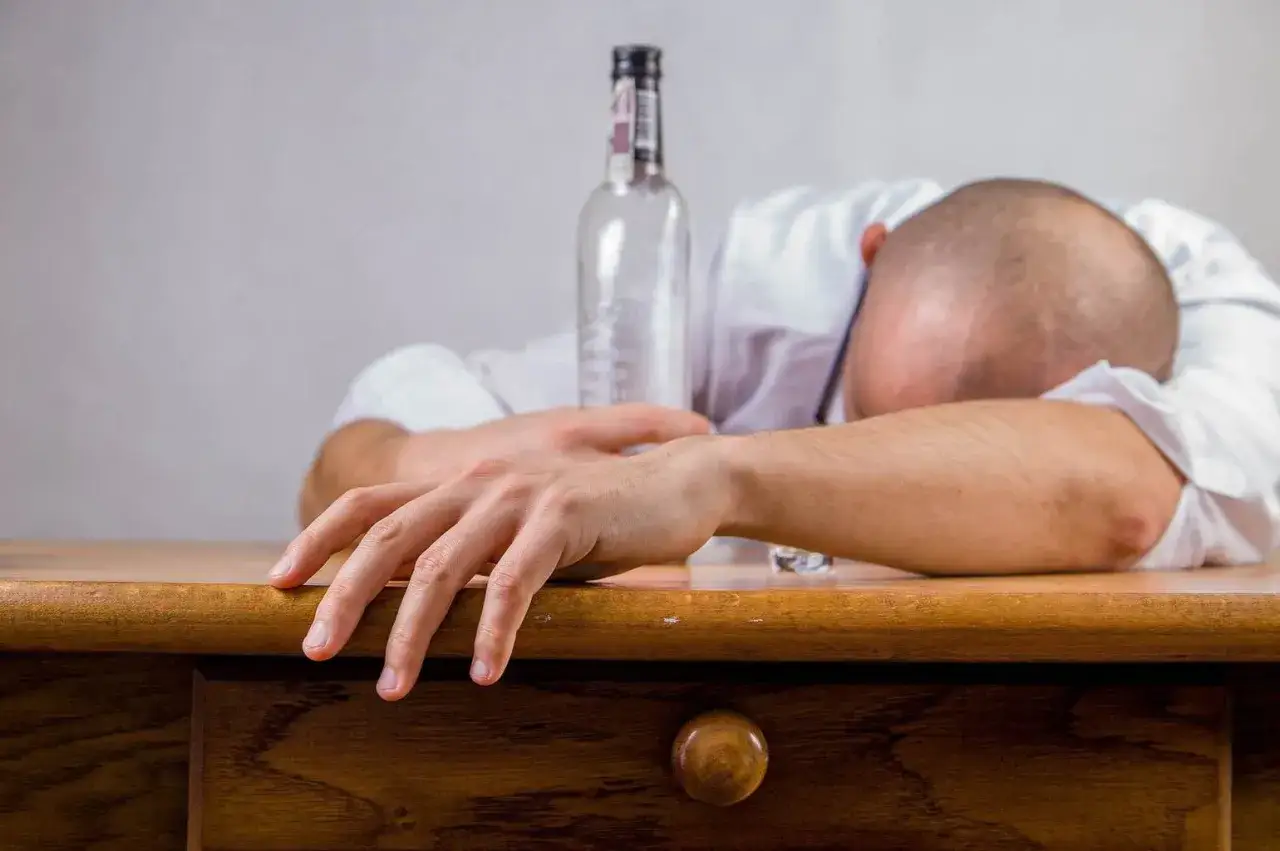 Mężczyzna śpi na biurku obok pustej butelki. To obrazuje problem alkoholowy, który może wpływać na ubezpieczenie PZU klauzula alkoholowa.