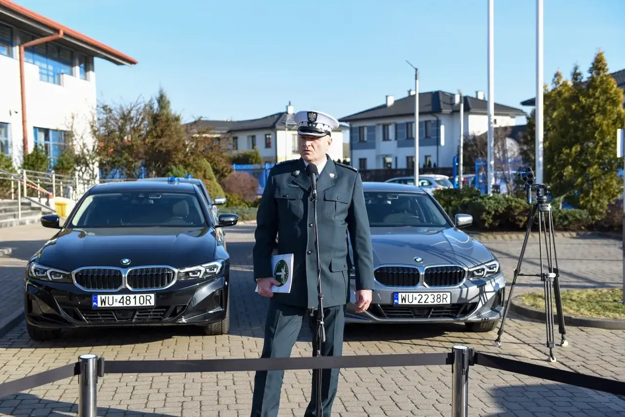 Funkcjonariusz w mundurze stoi przed dwoma nieoznakowanymi radiowozami.