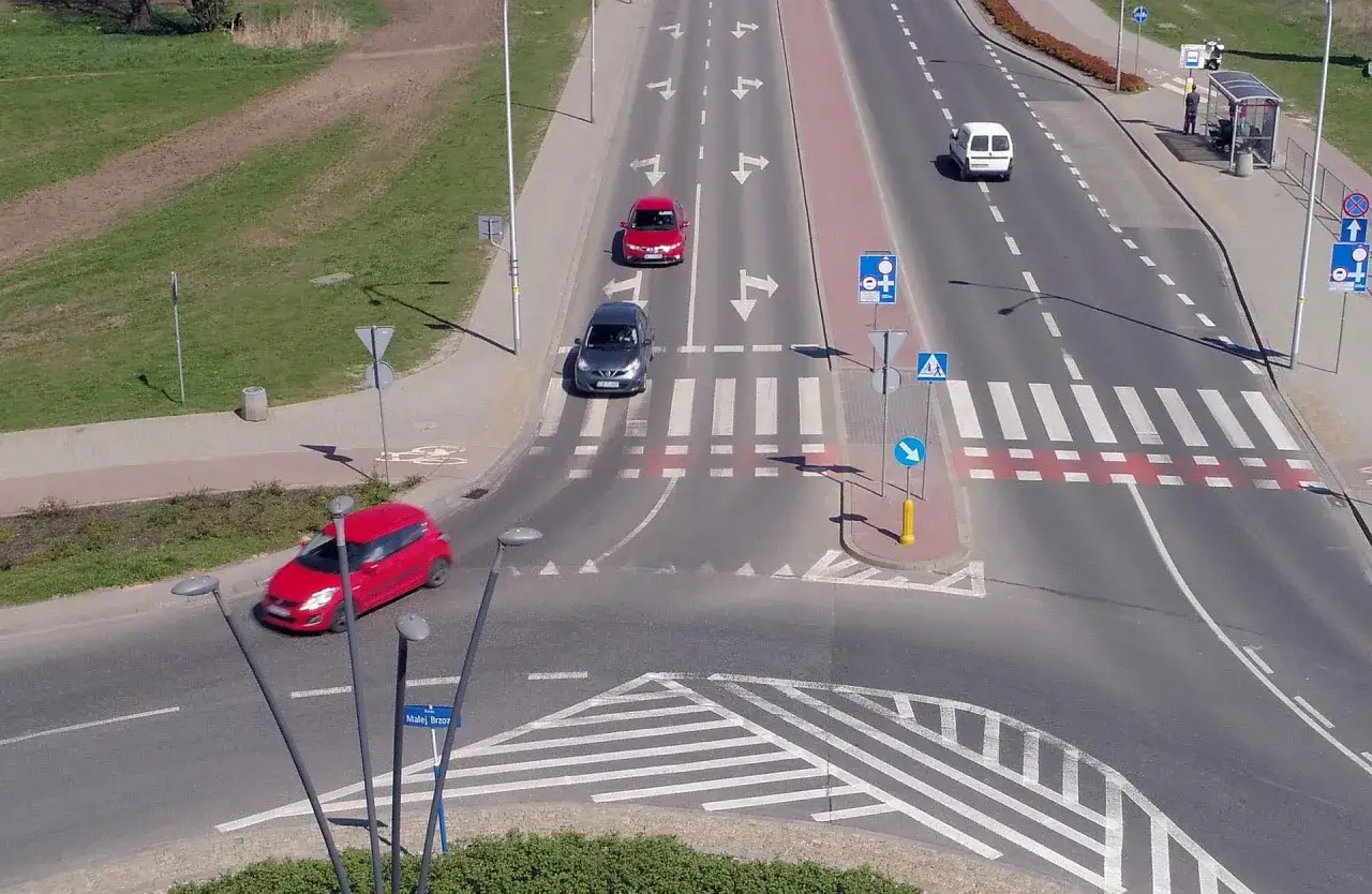 Na drodze widać różne rodzaje rond i przejść dla pieszych. Samochody poruszają się po pasach ruchu.